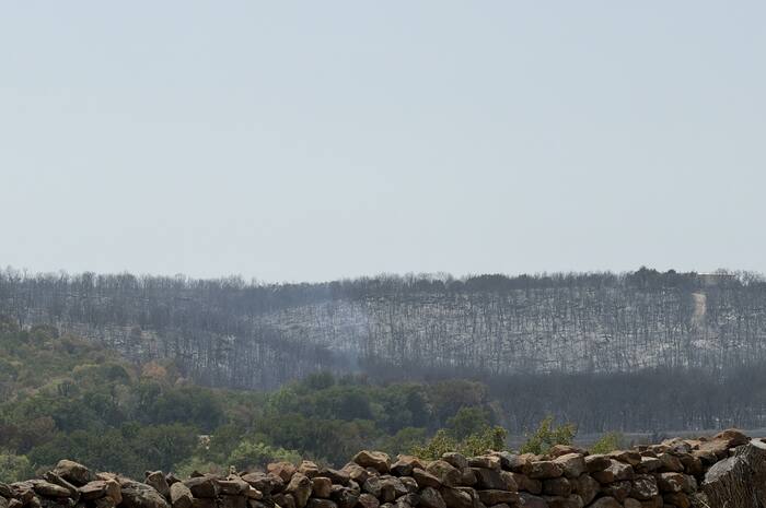 A stone wall overlooks a landscape with a forested hillside showing signs of recent fire damage, with smoke rising from the burned area.